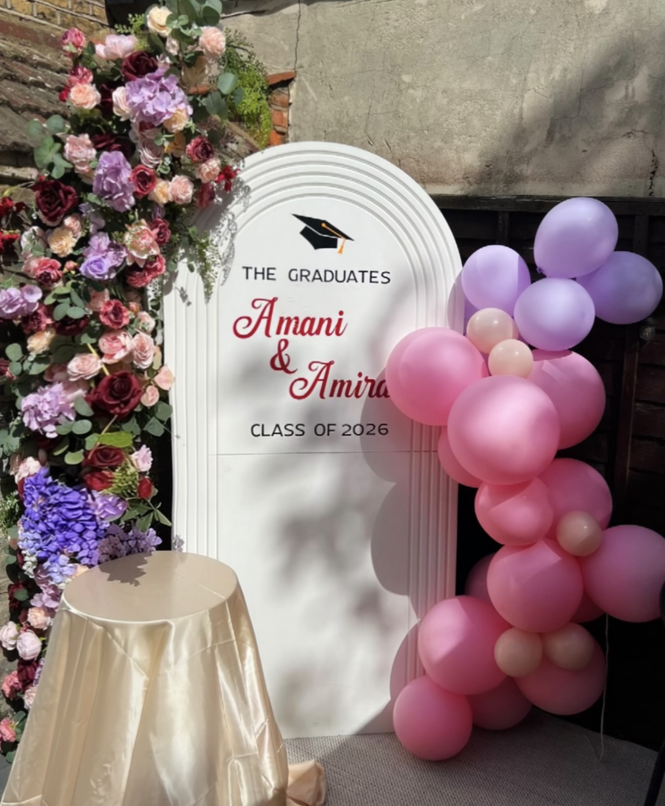 Personalised graduation board with a colourful flower arch, pink and purple balloons and a cake stand by Nour Occasions