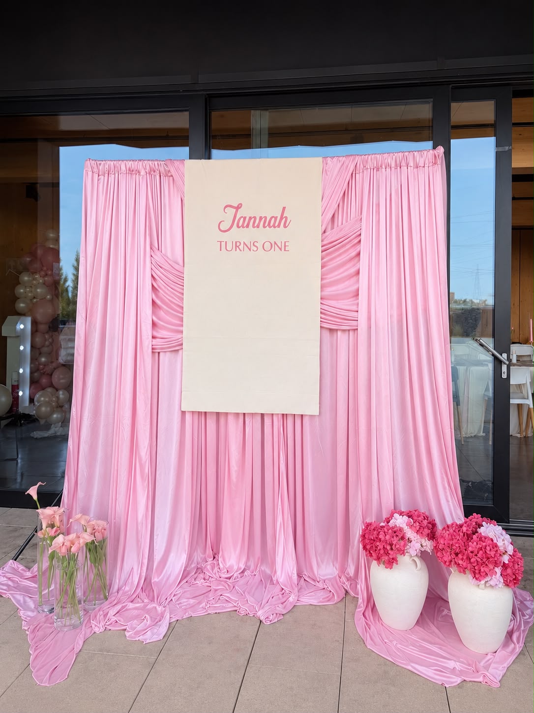 Pink first birthday draping with a personalised fabric banner and pink floral arrangements by Nour Occasions